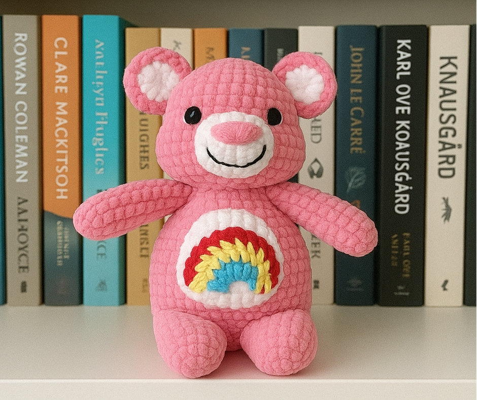 A crocheted amigurumi bear made of pink yarn, with a white snout, white inner ears, black safety eyes, embroidered black smile, and a white belly patch with a red, yellow, and blue rainbow. The amigurumi bear is sitting on a shelf in front of a row of books.