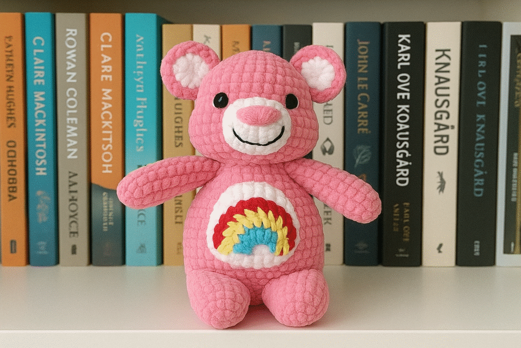 A crocheted amigurumi bear made of pink yarn, with a white snout, white inner ears, black safety eyes, embroidered black smile, and a white belly patch with a red, yellow, and blue rainbow. The amigurumi bear is sitting on a shelf in front of a row of books.