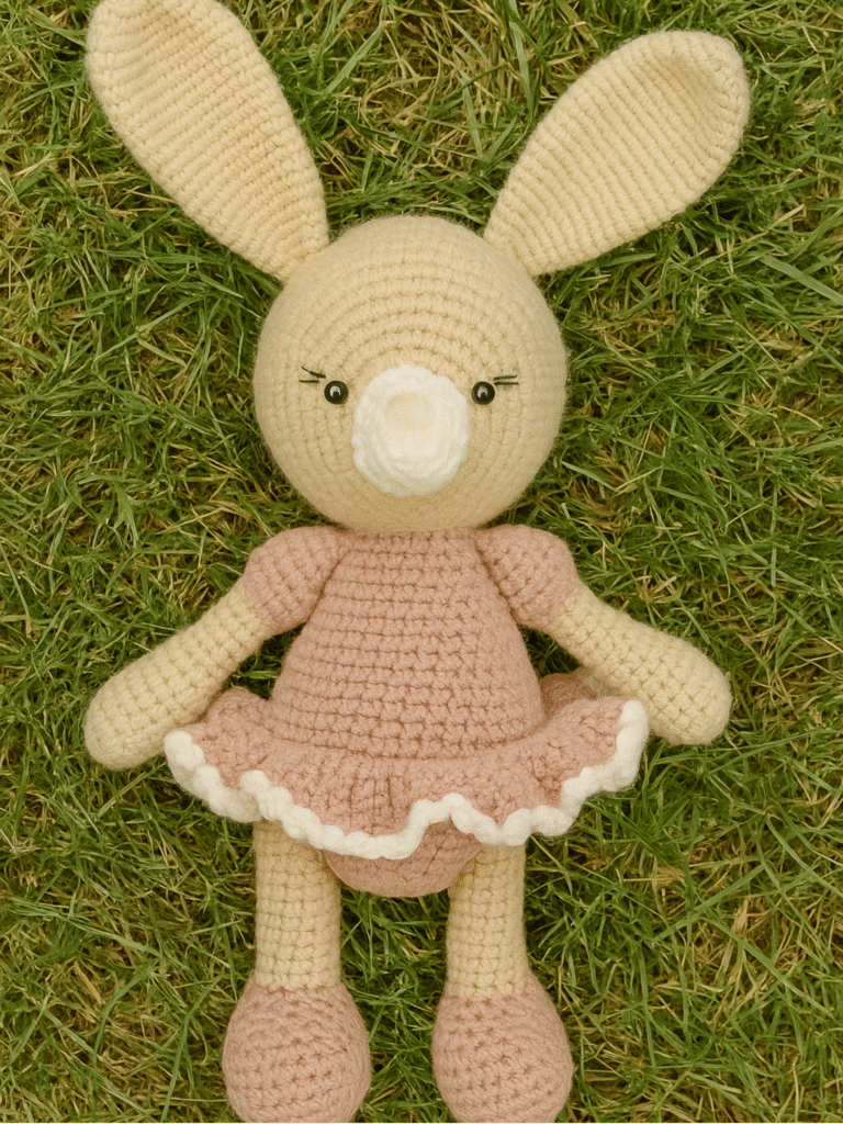 A handmade amigurumi bunny doll, primarily cream-colored with long ears, sits on green grass. It is wearing a light pink dress with a ruffled white trim. This adorable crochet creation is from LoopiaCrochet.