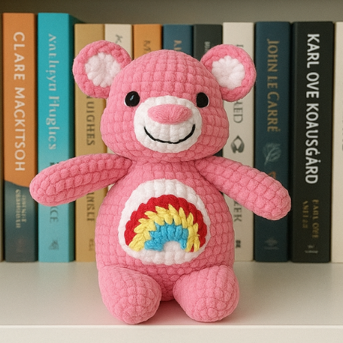 A pink crocheted amigurumi bear with a smiling face and a colorful rainbow design on its belly, sitting in front of a row of books. Free Amigurumi Bear Pattern.