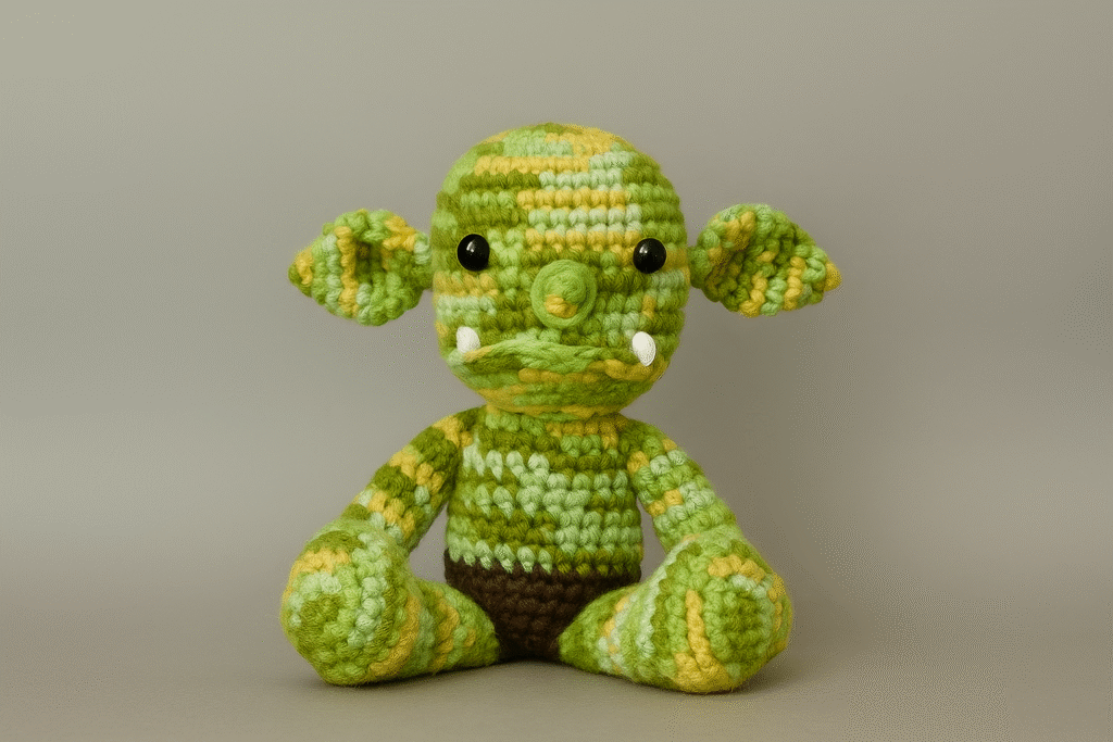 A handmade amigurumi ogre plush toy crocheted with variegated green and yellow yarn. The ogre has large, floppy ears, black button eyes, a round snout with two small white tusks, and wears dark brown crocheted shorts. It is posed sitting upright against a plain gray background.