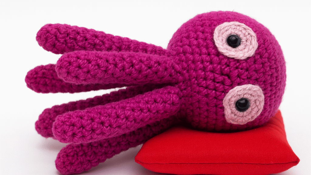 A purple amigurumi octopus lying on a red fabric pillow. The octopus has two large round eyes with black pupils and pink eyelids. The text on the image suggests a free octopus crochet pattern.