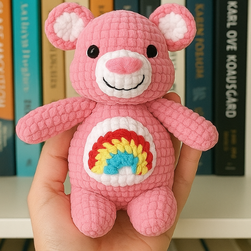 A hand holds up a crocheted pink teddy bear with a rainbow on its belly.