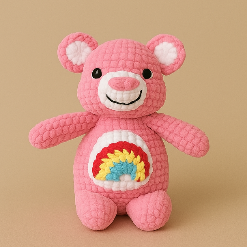A small, crocheted pink teddy bear with a rainbow on its belly sits against a solid tan background.