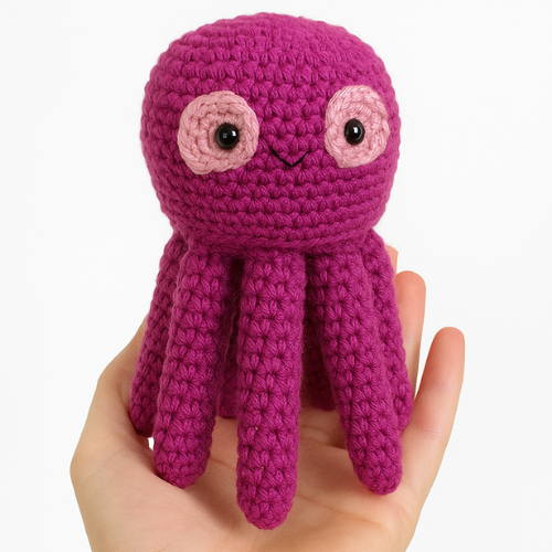 A completed purple amigurumi octopus with eight tentacles, held in someone's hand, representing a free octopus crochet pattern. The octopus has two large pink-rimmed eyes and a small stitched smile.