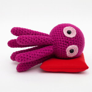 A cute, purple crocheted octopus with large, pink eyes and black pupils rests on a small red pillow. Keyword: Free Octopus crochet pattern.