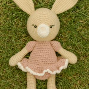 A crocheted amigurumi bunny doll with long ears and a light-colored body, wearing a pastel pink dress with a ruffled white trim, lying on a patch of green grass. The doll's facial features are embroidered, and its nose is a small white pom-pom. The image is for an Adorable Bunny Crochet Pattern PDF.