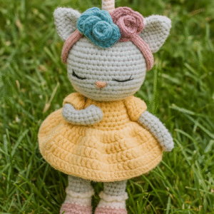 A handcrafted amigurumi unicorn doll with closed, embroidered eyes and eyelashes, a silver-gray head, and a pink horn. It is wearing a pink headband with three crocheted flowers in teal and pink, a ruffled yellow dress, and has a pink nose. The doll is standing on a patch of green grass.