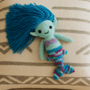 An amigurumi doll of a mermaid with a turquoise head and torso, purple seashell top, and a striped blue, purple, and turquoise tail. Her long hair is made of bright blue yarn strands. The doll is lying on a pillow with a striped pattern.
