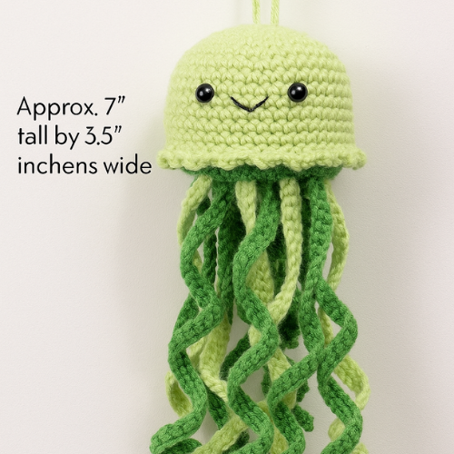 A crocheted green jellyfish toy with black eyes and a smile. Text indicates its approximate size: 7" tall by 3.5" wide.