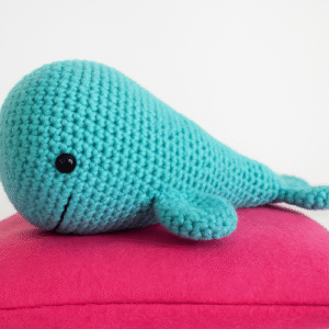 A small, turquoise blue crocheted whale sits on a bright pink cushion. The whale has a simple, stitched smile and a black bead for an eye.