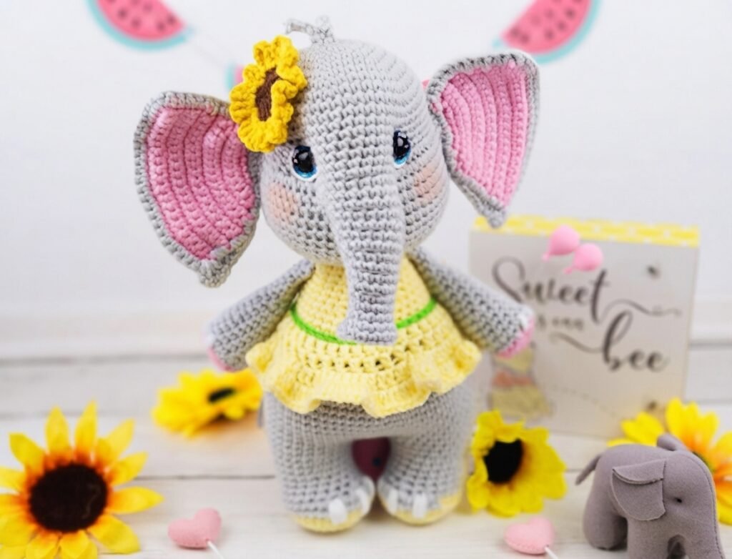 A handmade amigurumi elephant crochet toy featuring grey yarn, big pink-lined ears, a yellow dress, and a small yellow sunflower attached to its head, standing on a white wooden surface with decorative sunflowers and hearts around it.