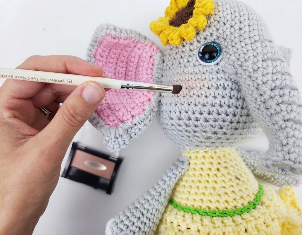 A hand applies blush with a brush to a crocheted amigurumi elephant toy as part of a free elephant crochet pattern tutorial.