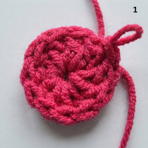 A close-up photograph of a crocheted circle made from hot pink yarn. The circle is centered on a plain white background. The texture of the crochet stitches is clearly visible, radiating from the center point which has a visible opening (the starting magic ring). A short loop of pink yarn and a long, trailing yarn tail are visible at the top right of the circle, indicating it is a work-in-progress. The number "1" is printed in black in the top right corner of the image.