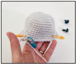 A hand holding a light gray crocheted amigurumi piece marked with yellow and blue stitch markers, with two safety eyes placed beside it on a white background.