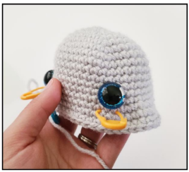 A hand holding a light gray crocheted amigurumi piece with two safety eyes inserted into the sides and a yellow stitch marker attached to the top edge.