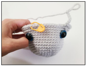 A hand holding a light gray crocheted amigurumi piece with a large blue safety eye and a yellow stitch marker placed below it.   