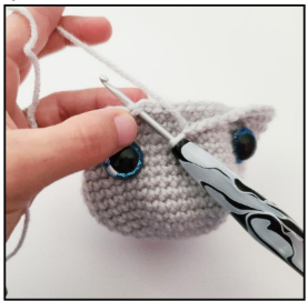 A hand holding a light gray crocheted amigurumi piece with a large blue safety eye and a yellow stitch marker placed below it.   