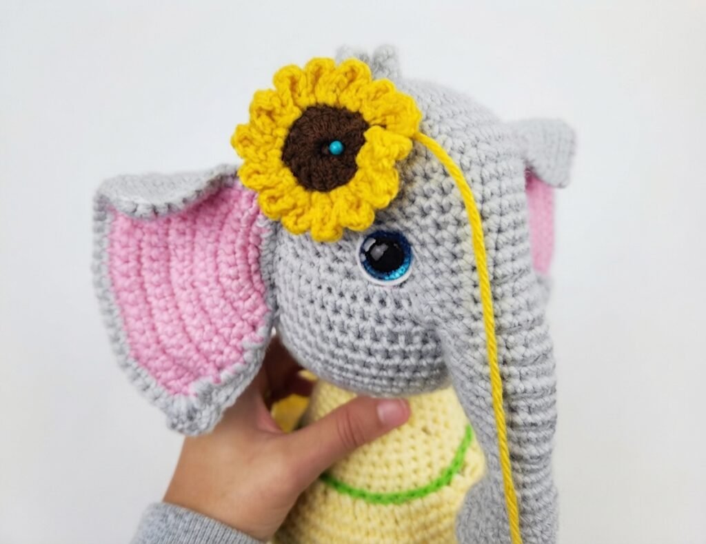 A handmade grey amigurumi elephant crochet toy with pink-lined ears, a yellow dress, and a small yellow sunflower accessory standing on a white wooden surface with decorative sunflowers and hearts, featured in a free elephant crochet pattern tutorial.