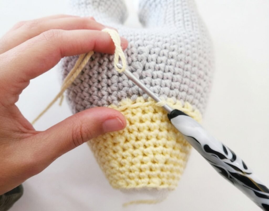 A close-up view of a hand holding a crochet hook to attach yellow ruffles onto the grey body of a handmade amigurumi elephant, as part of a free elephant crochet pattern tutorial.
