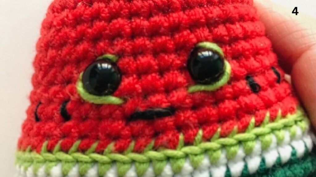Step 4 of a free watermelon keychain crochet pattern, showing a close-up of the crocheted watermelon face with embroidered black seed details, safety eyes outlined with light green yarn, a stitched black mouth, and the number '4' in the upper corner, all against a light beige background.