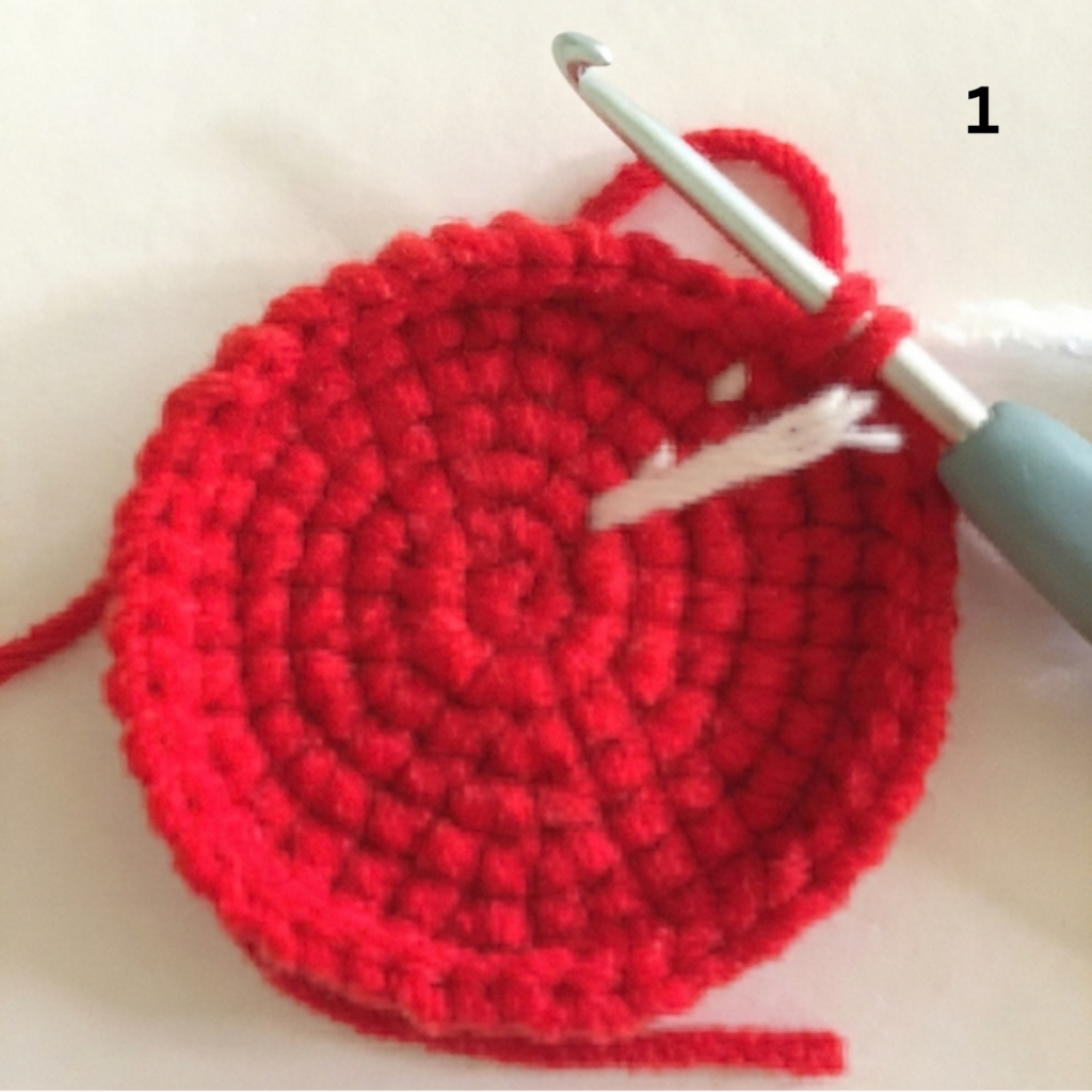 Step 1 of making a crochet watermelon slice from a keychain free crochet pattern, showing a round red piece worked in single crochet with a crochet hook and stitch marker on a light beige background