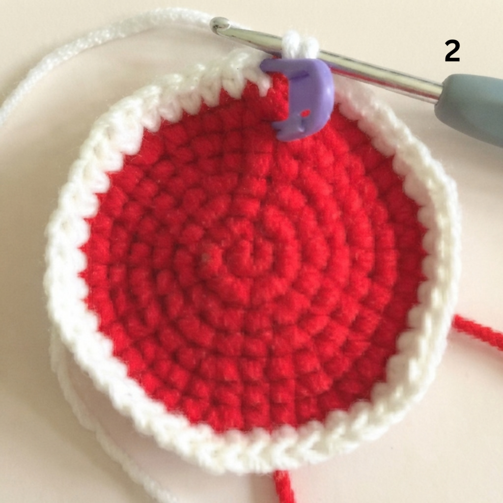 Step 2 of making a crochet watermelon slice from a free keychain crochet pattern, showing a round red piece edged with white single crochet, a purple stitch marker, and a crochet hook on a light beige background