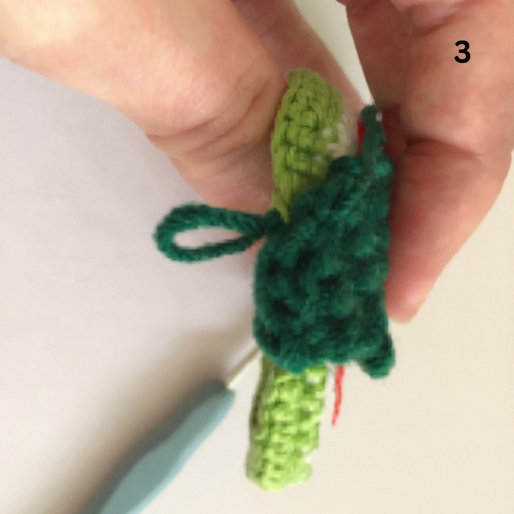 Step 3 of making a crochet watermelon slice from a free keychain crochet pattern, showing a hand holding the folded green and red crocheted piece with a hanging loop on a light beige background
