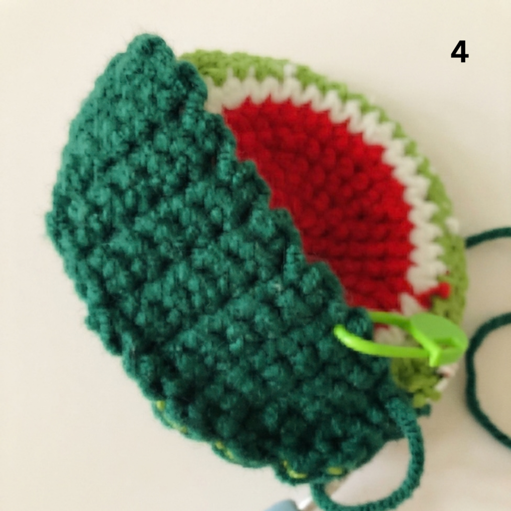 Step 4 of assembling a crochet watermelon slice keychain, following a free keychain crochet pattern, showing the dark green outer layer being attached to the inner red, white, and light green piece using a green stitch marker and a crochet hook, all on a light beige background