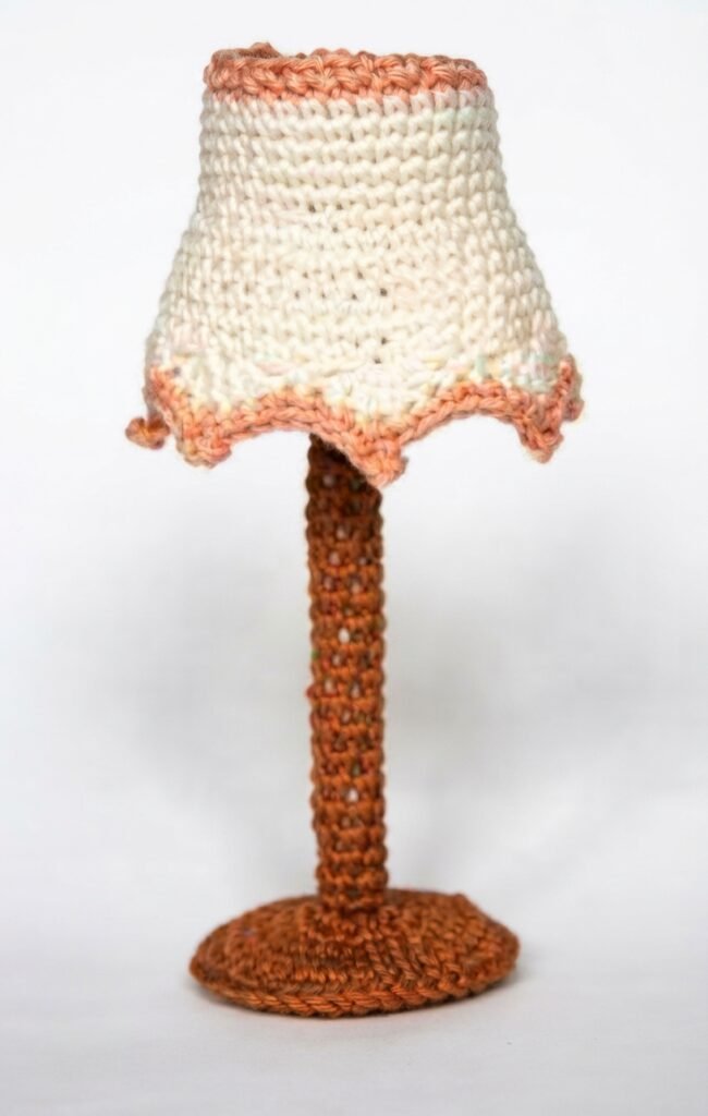 A high-resolution, detailed photograph of a miniature, hand-crocheted table lamp featuring a brown stem, a rounded base, and a cream-colored lampshade trimmed with scalloped orange edging. This miniature lighting accessory for a Crochet Mini Living Room stands upright against a seamless white background.