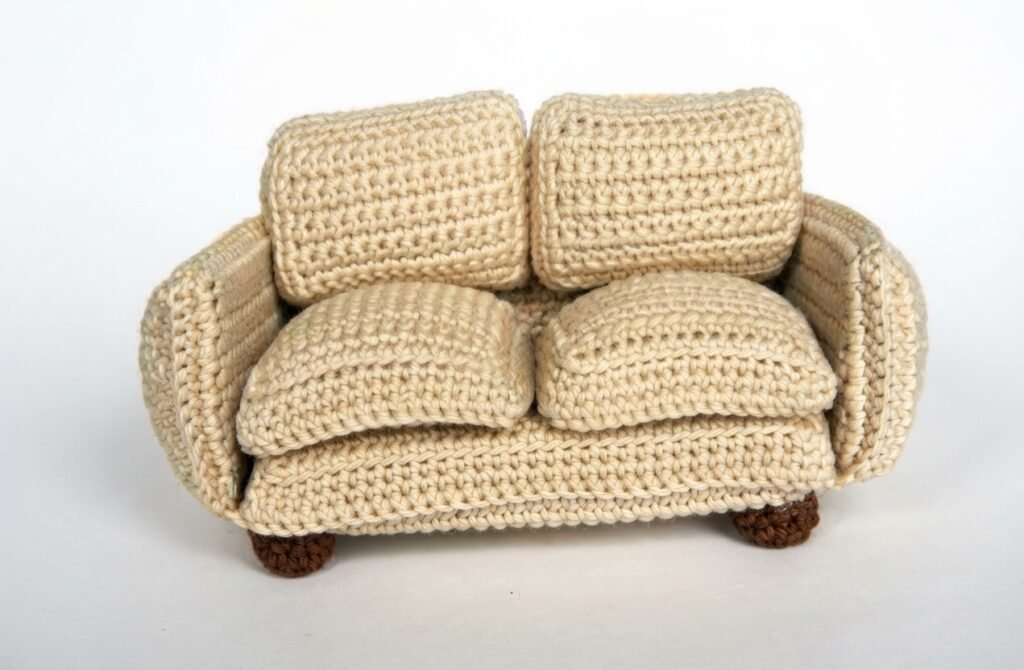A high-resolution, detailed photograph of a miniature, hand-crocheted two-seater sofa, crafted from light beige yarn with a distinct single-crochet texture. The miniature furniture for a Crochet Mini Living Room features two seat cushions and two back cushions, and stands on four small, textured brown bun-feet against a seamless white background.