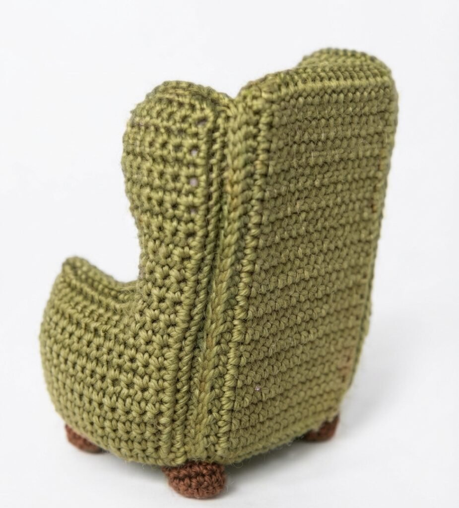 A high-resolution, detailed photograph of the back and three-quarter view of the miniature moss-green crocheted armchair, featuring a prominent vertical seam detail. This miniature, hand-stitched piece for a Crochet Mini Living Room stands on four small, textured brown bun-feet against a seamless white background.