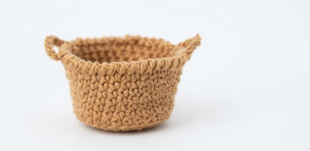 A high-resolution, detailed photograph of a miniature, hand-crocheted storage basket in a warm tan color, featuring a textured single-crochet stitch and two small, integrated loop handles[cite: 15]. This miniature decor accessory for a Crochet Mini Living Room sits against a seamless white background