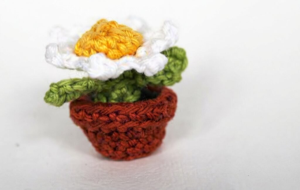 A high-resolution, detailed photograph of a miniature, hand-crocheted potted flower in a reddish-brown clay pot, featuring green leaves and a white daisy with a bright yellow center. This charming miniature decor piece for a Crochet Mini Living Room sits against a seamless white background.