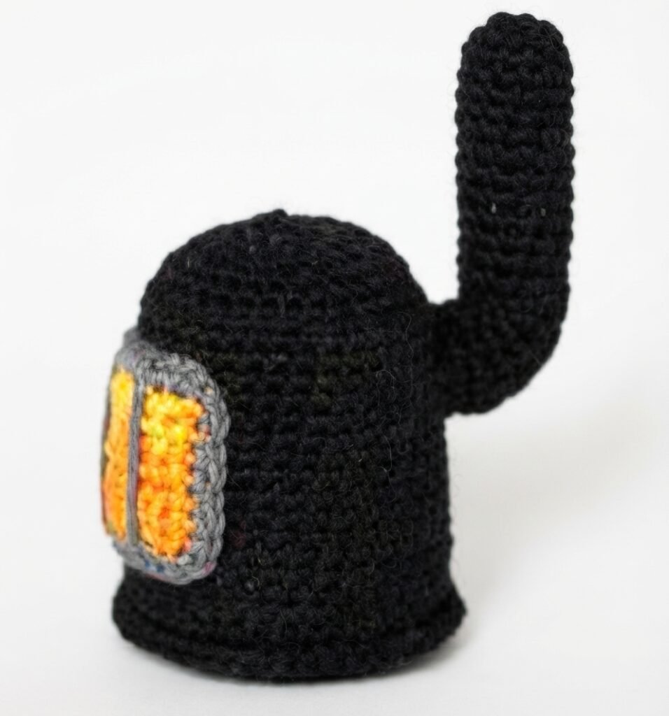 A high-resolution, detailed photograph of a hand-crocheted black crewmate figurine from the game Among Us, featuring a grey-framed visor with glowing orange-yellow texture and an upright antenna. This miniature character piece for a Crochet Mini Living Room stands against a seamless white background.