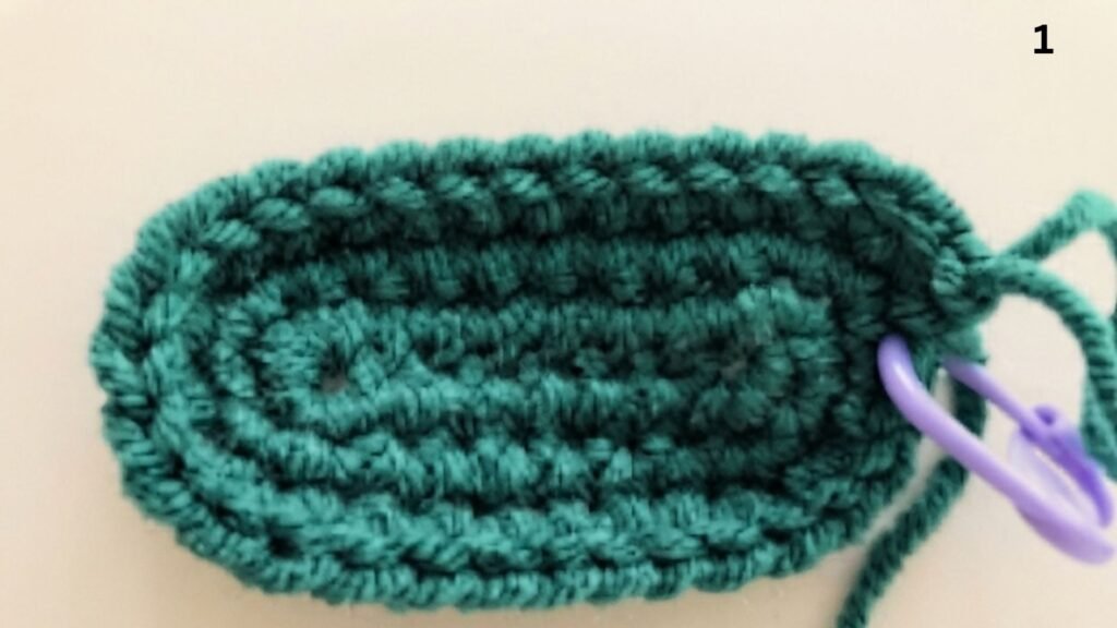 Step 1 of a free keychain crochet pattern, showing an oval-shaped base made with dark teal single crochet stitches, with a purple stitch marker attached to the last stitch and the number '1' in the corner, all on a light beige background.