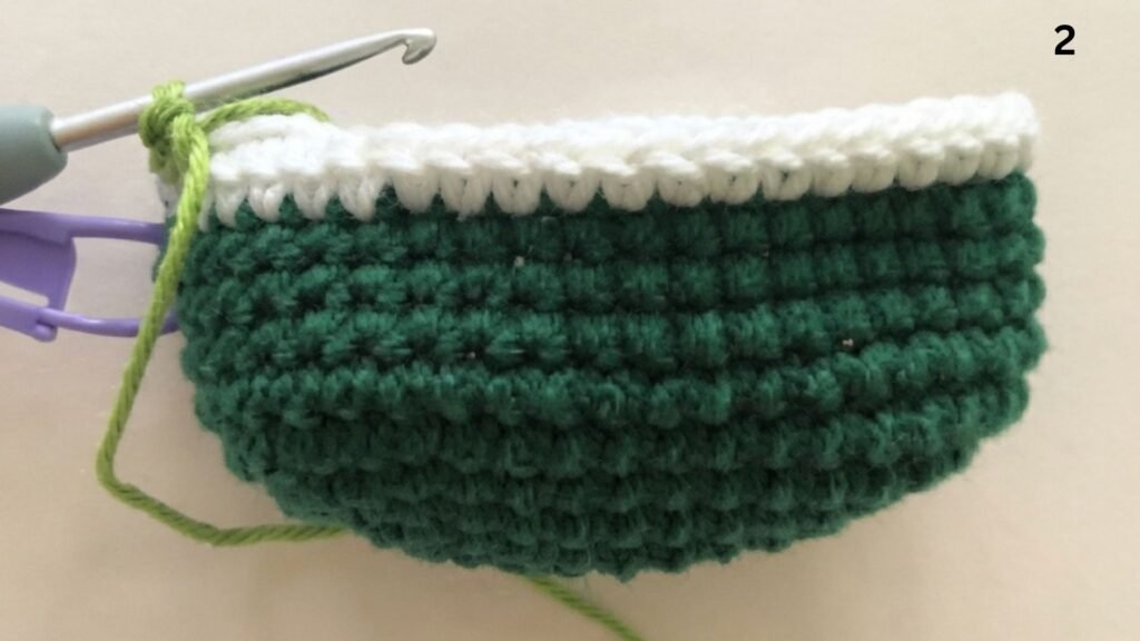 Step 2 of a free watermelon keychain crochet pattern, showing the work-in-progress piece with dark green and white rows, a light green yarn loop on a crochet hook, a purple stitch marker, and the number '2' in the top right corner, all set against a light beige background.