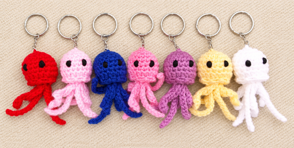 Free Keychain Crochet pattern featuring seven colorful handmade octopus keychains arranged in a row on a light beige background, each with soft yarn texture, black eyes, and silver key rings